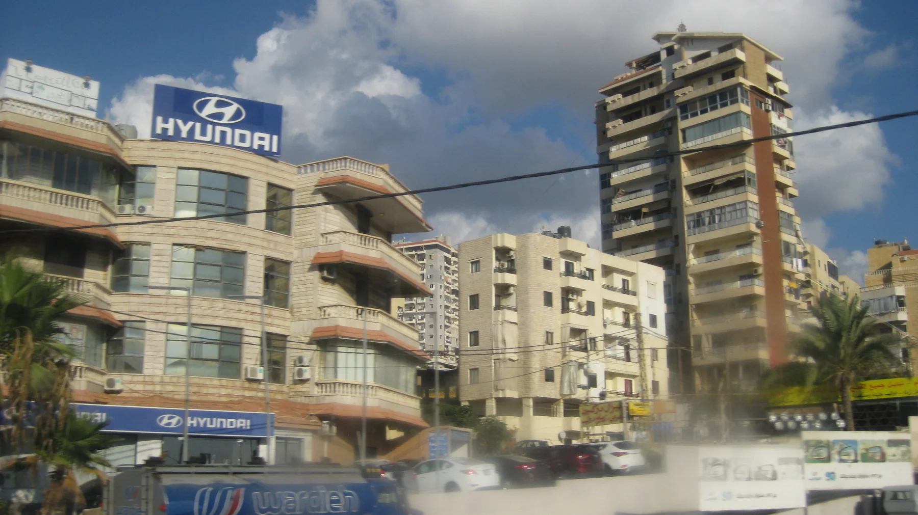 Buildings with balconies, commercial signs and a tall narrow apartment block stand behind a busy roadway.