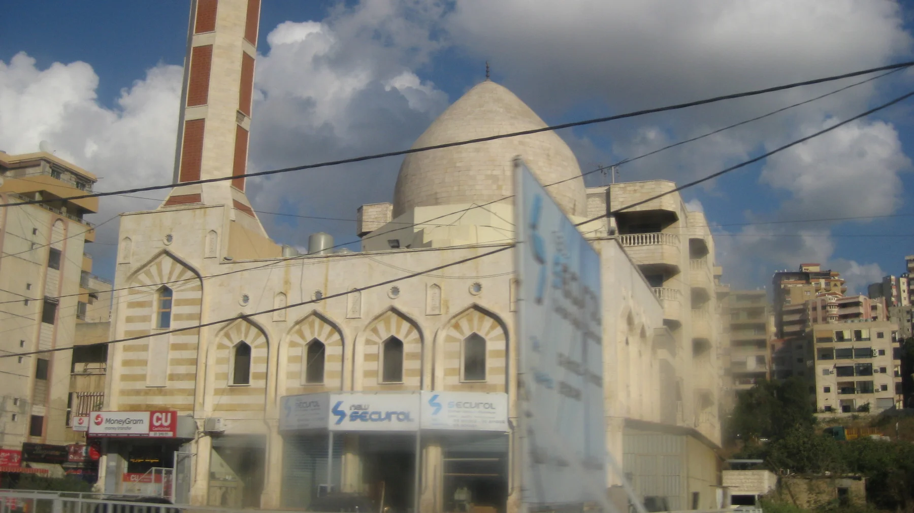 A domed building with arched windows and a tall striped minaret stands among urban blocks and overhead wires.