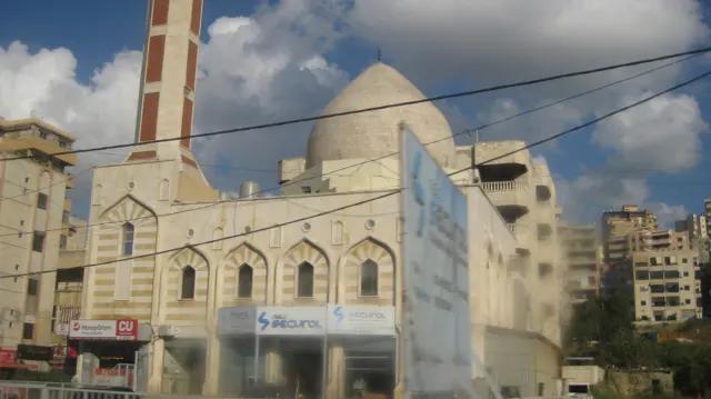 A domed building with arched windows and a tall striped minaret stands among urban blocks and overhead wires.