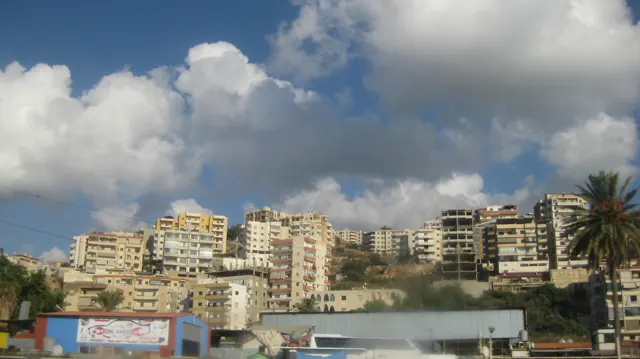Rows of pale apartment buildings cover a slope beneath bright clouds, with roadside signs below.