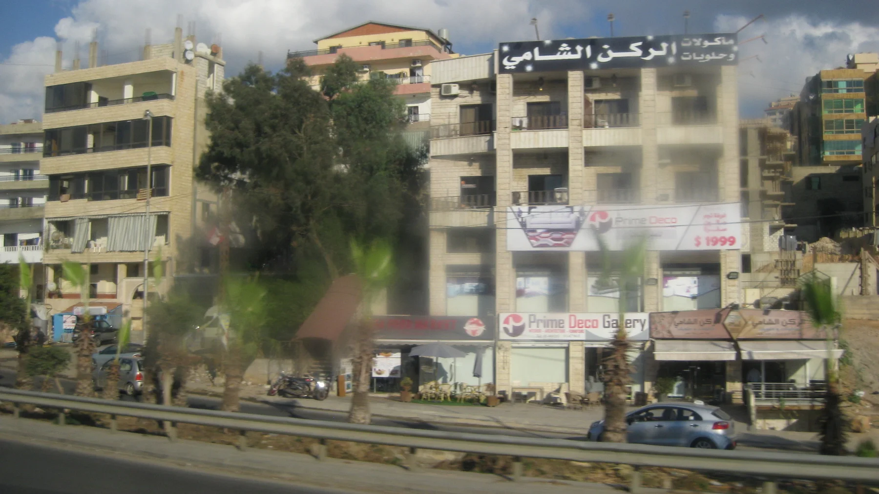 Cars pass buildings with balconies and shop signs beside a central roadside barrier.