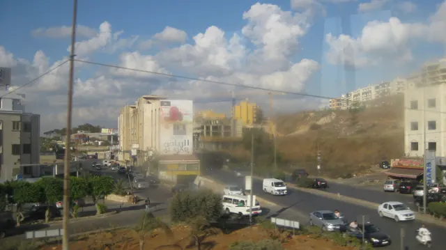 Vehicles follow a broad divided road between buildings and a dry hillside beneath scattered clouds.