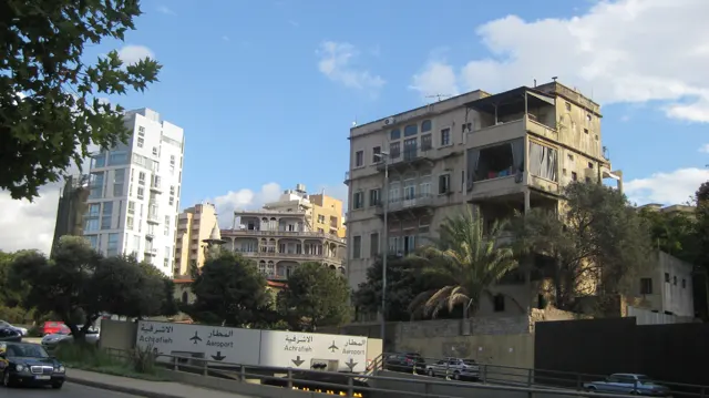 Older apartment blocks and a tall white building stand behind trees and a low roadside fence.