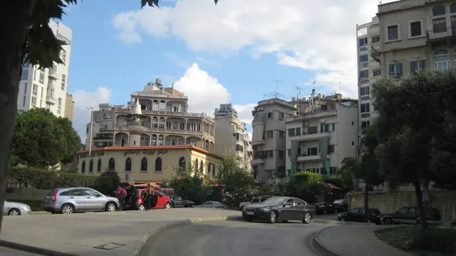 Cars pass an open junction surrounded by buildings with balconies, trees and a yellow corner facade.