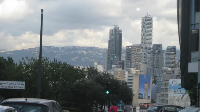 Tall modern towers rise beyond trees and a green traffic signal, with hills in the distance.