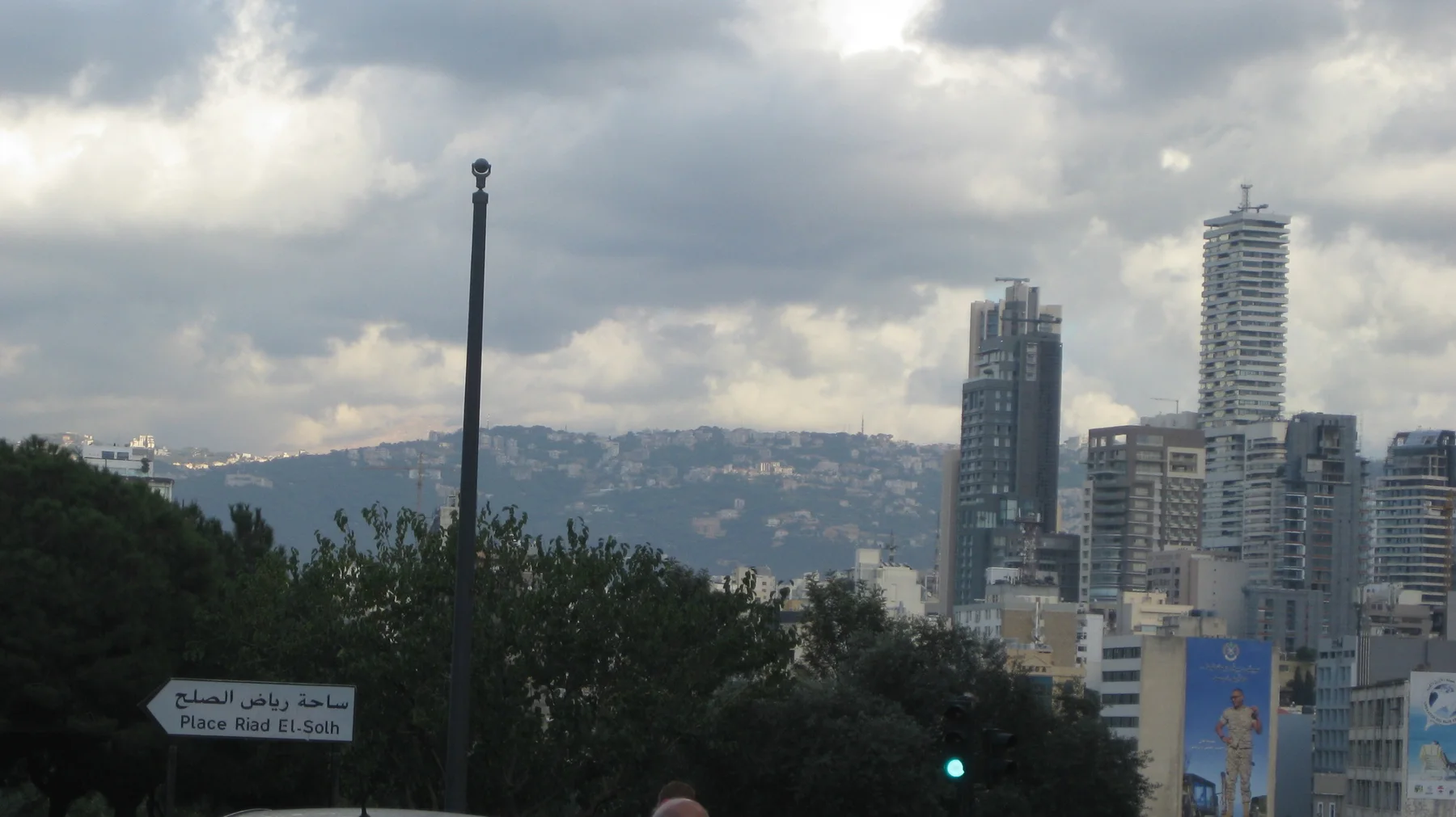 A cluster of high-rises stands before distant hills beneath layered clouds, partly screened by trees.