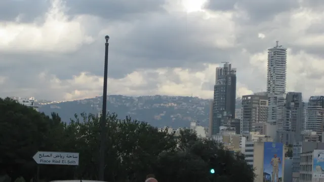 A cluster of high-rises stands before distant hills beneath layered clouds, partly screened by trees.
