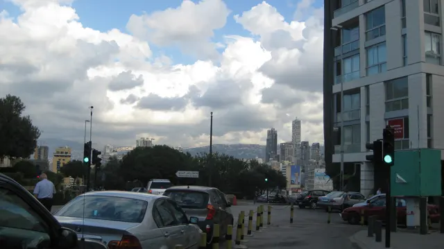 Traffic gathers at a junction beside a tall building and green lights beneath a clouded sky.
