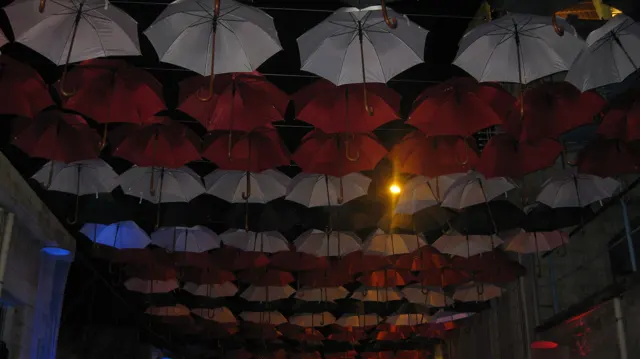 Rows of red and pale umbrellas hang above a narrow street, lit by scattered lamps after dark.