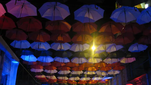 Suspended umbrellas glow red, blue and purple over a brightly lit night-time passage.