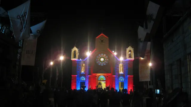 A church facade with a circular window and small towers glows in red and blue lighting at night.
