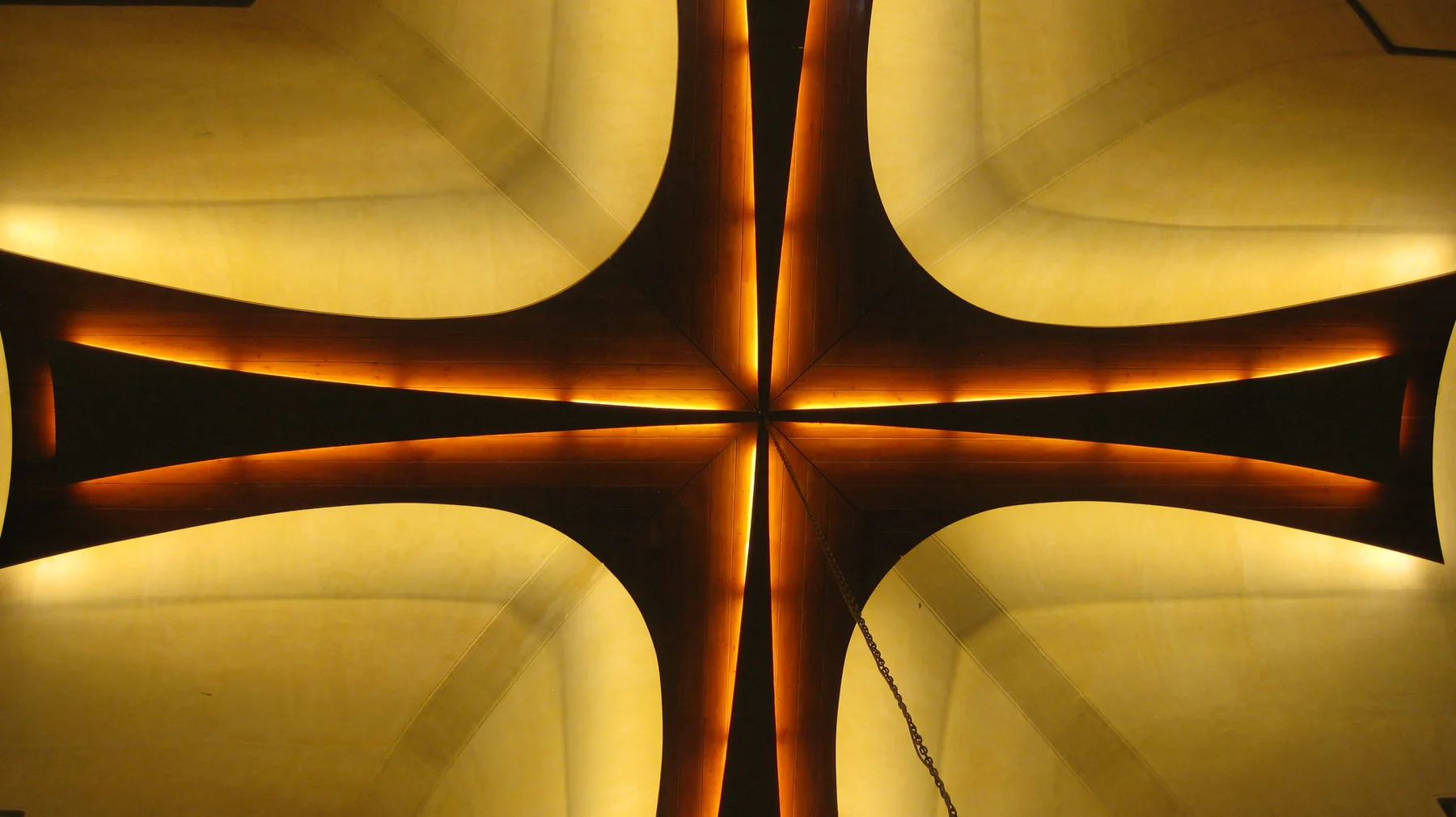 A dark cross-shaped structure is outlined by warm golden lighting against a pale curved background.