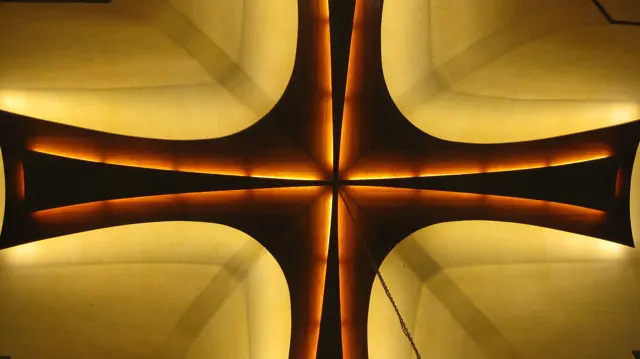 A dark cross-shaped structure is outlined by warm golden lighting against a pale curved background.