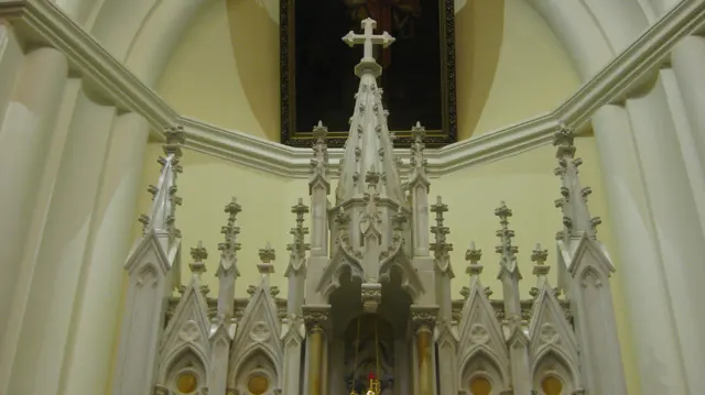 A richly decorated white altar with pointed spires and a cross stands beneath a pale church arch.