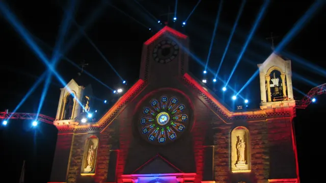Blue light beams fan into the night sky above a church facade outlined in red and warm light.