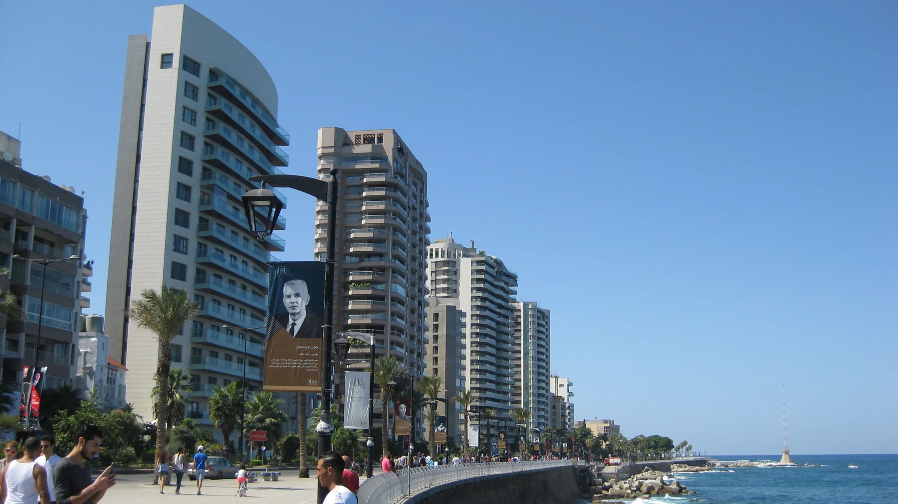 People walk beside a line of high-rise buildings and palm trees along a broad seafront promenade.