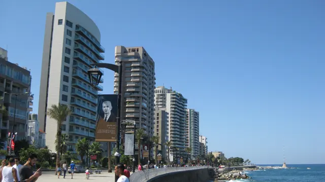 People walk beside a line of high-rise buildings and palm trees along a broad seafront promenade.