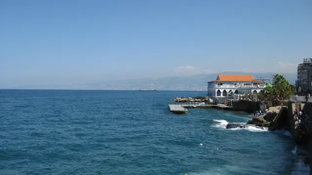 A white waterfront building with an orange roof stands beside blue water and a rocky shoreline.