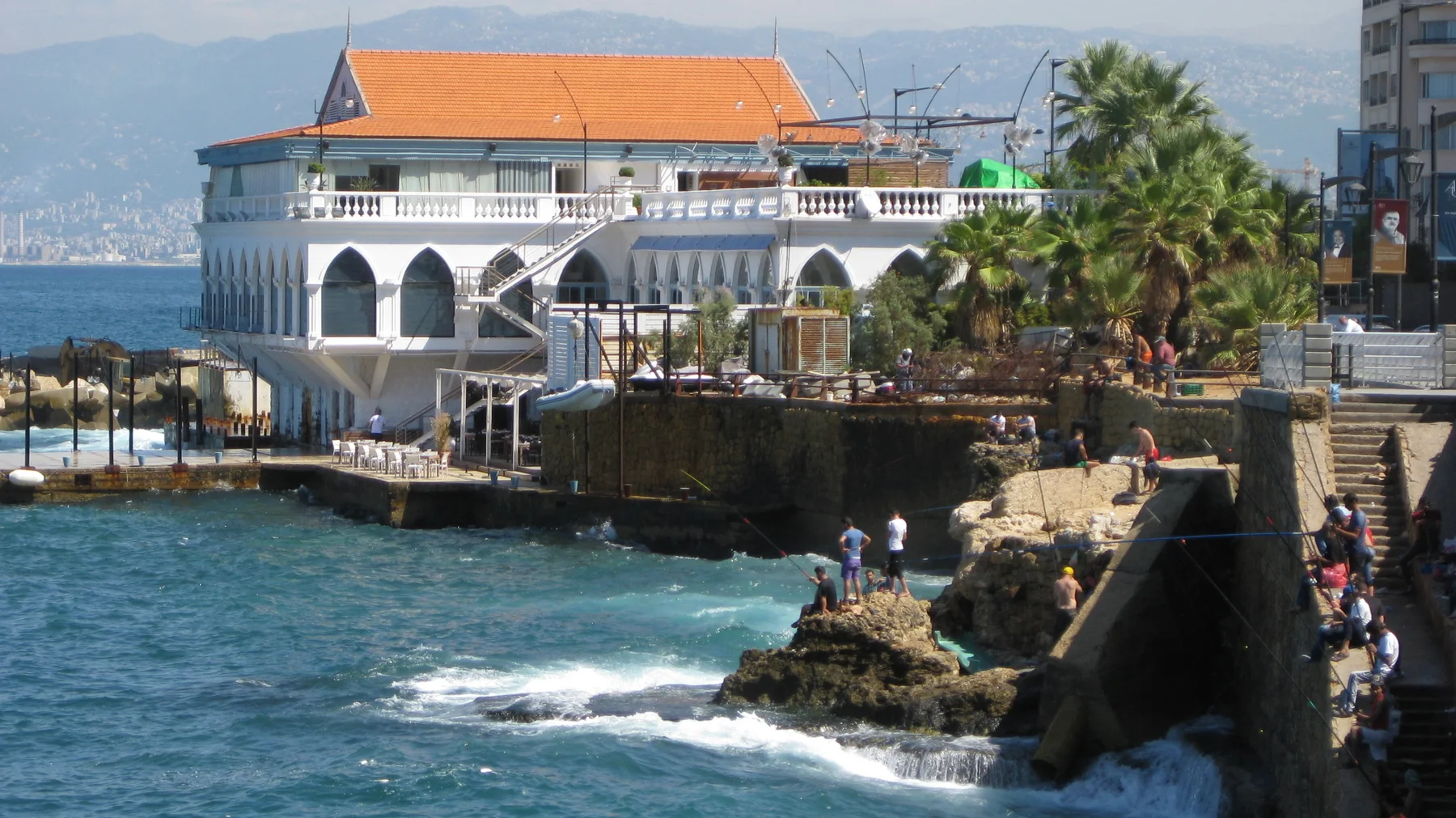Waves break beneath a waterfront building with white arches, an orange roof and palm-lined terraces.