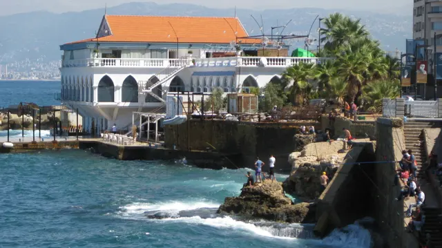 Waves break beneath a waterfront building with white arches, an orange roof and palm-lined terraces.
