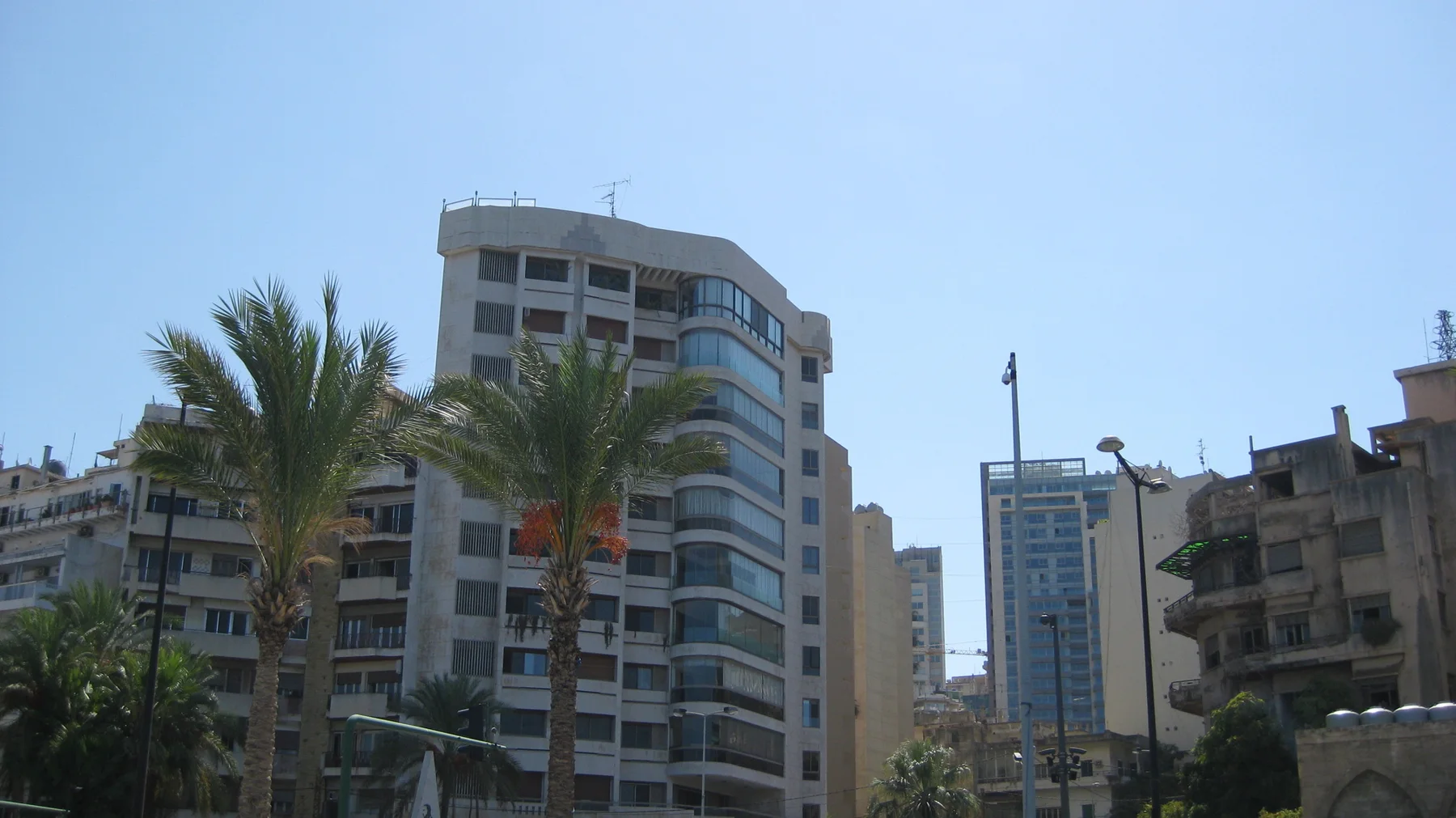 Palm trees rise in front of apartment buildings with balconies beneath a clear blue sky.