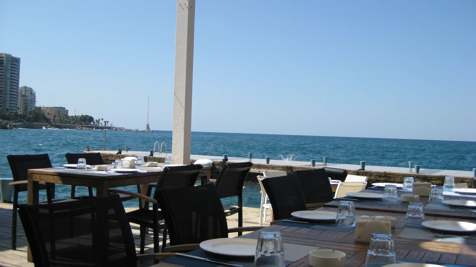 Outdoor tables set with plates and glasses face the blue sea beneath a shaded terrace roof.