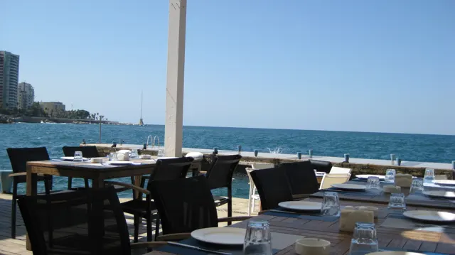 Outdoor tables set with plates and glasses face the blue sea beneath a shaded terrace roof.