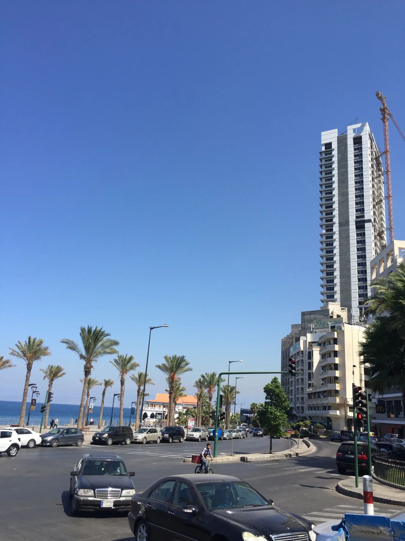 A tall narrow building rises above traffic at a palm-lined junction near the sea.