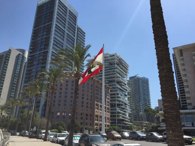 A Lebanese flag hangs among palms and high-rise buildings above street traffic.