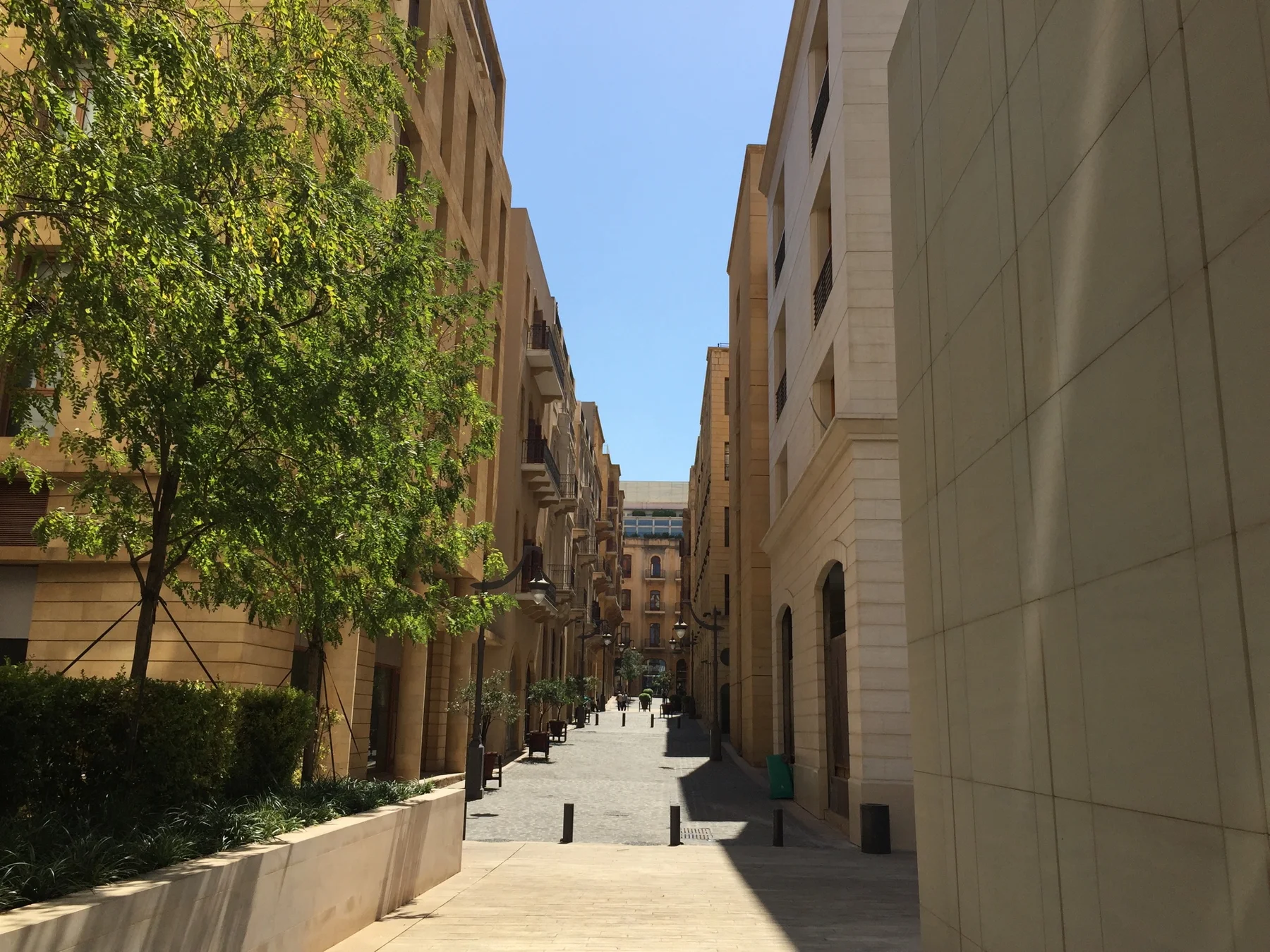 A narrow sunlit pedestrian passage runs between pale buildings beside trees and low planting.