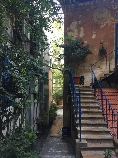 A narrow stairway rises beside a reddish building, dark railings and dense green climbing plants.