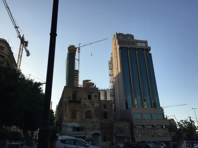 Tall cranes and a modern tower rise above lower buildings in fading daylight.