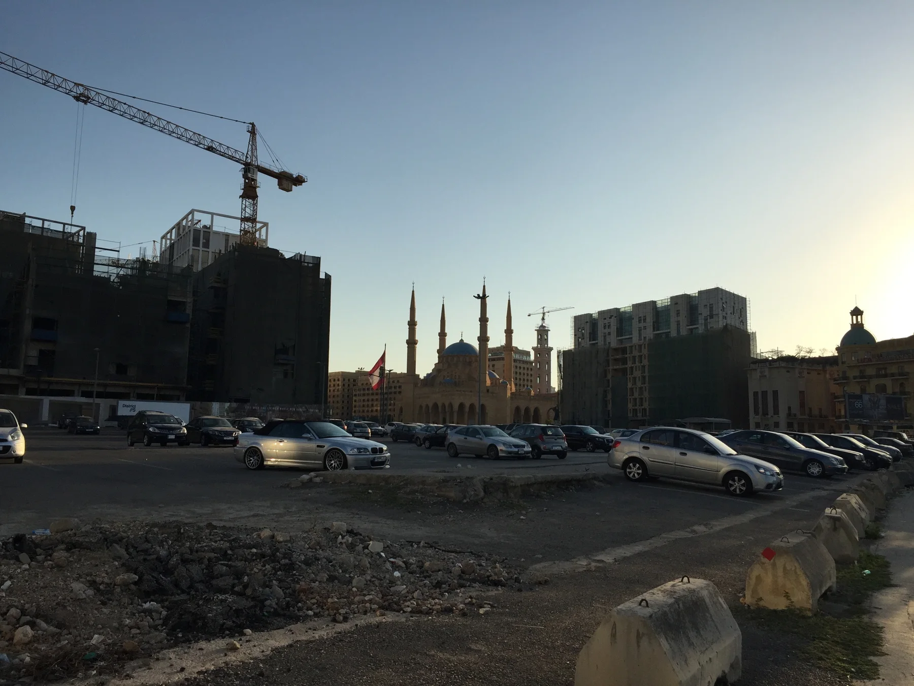 Rows of cars sit before distant minarets and a domed building beneath a construction crane.