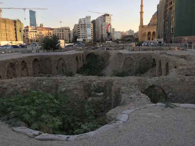 Low excavated stone walls and patches of greenery lie below surrounding buildings and a distant minaret.