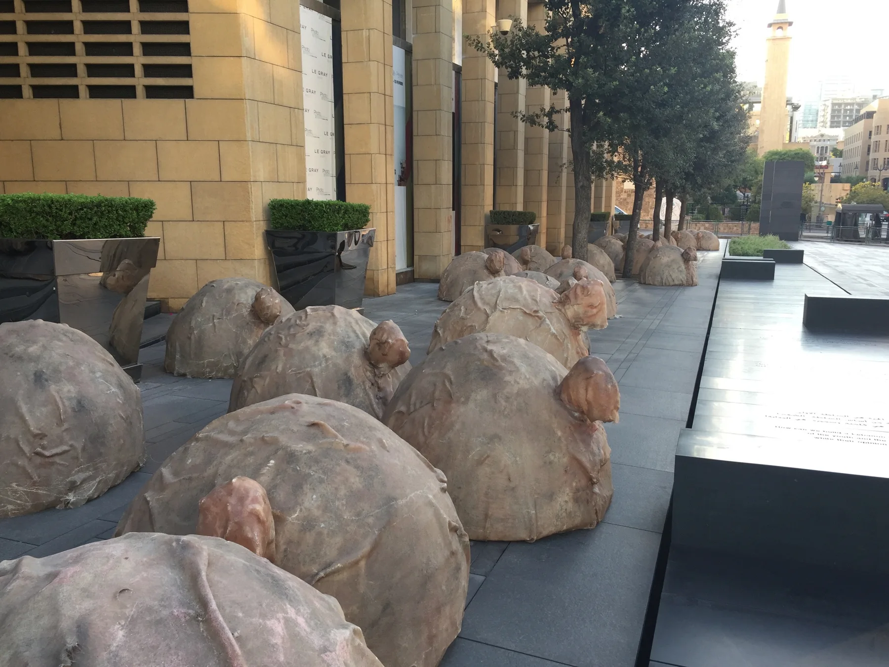 Rounded sculptural stone forms sit on low platforms beside a paved urban square and pale buildings.
