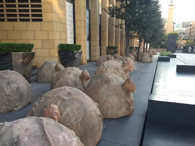 Rounded sculptural stone forms sit on low platforms beside a paved urban square and pale buildings.