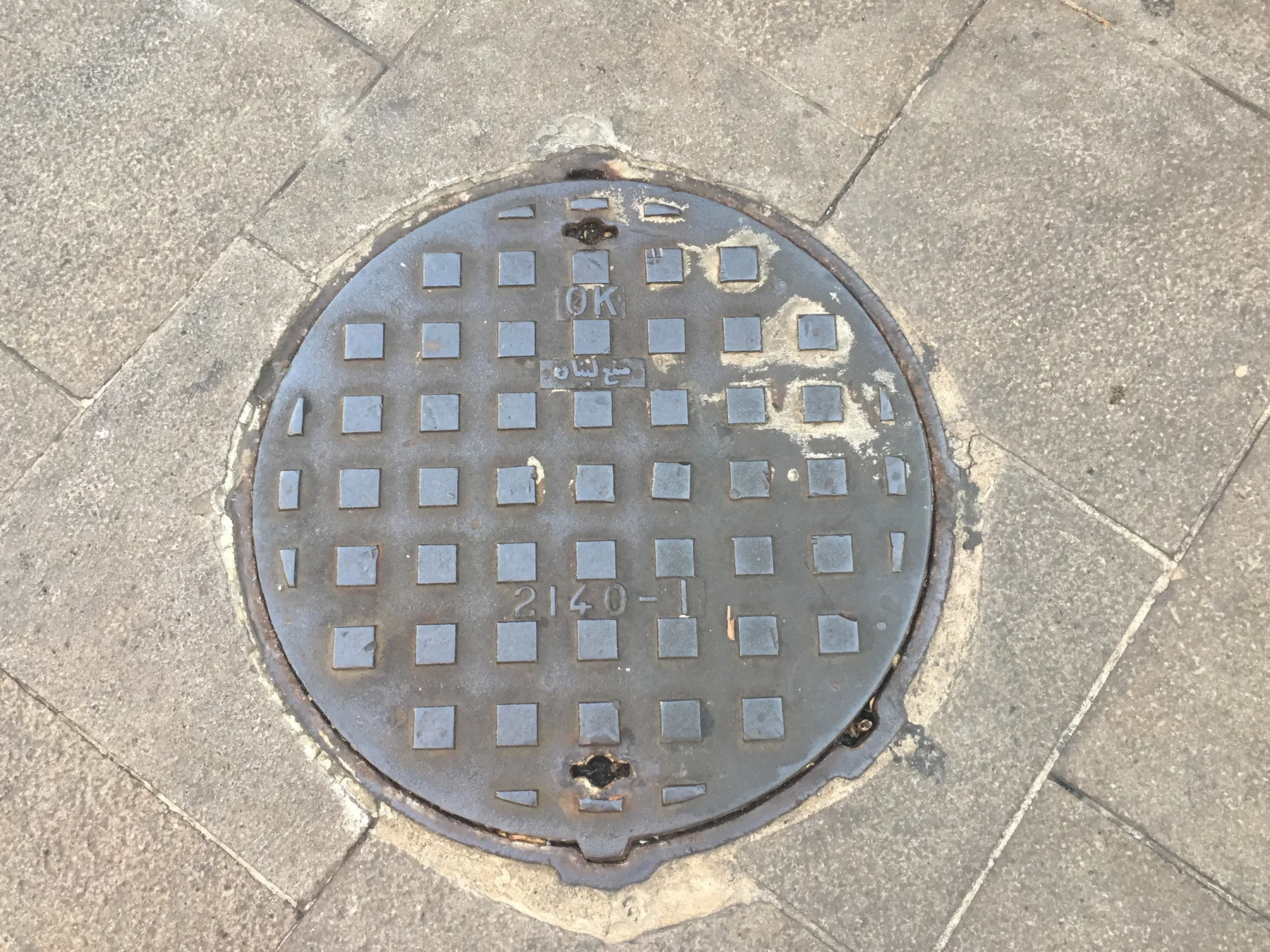 A round utility cover patterned with small squares sits within pale stone paving.