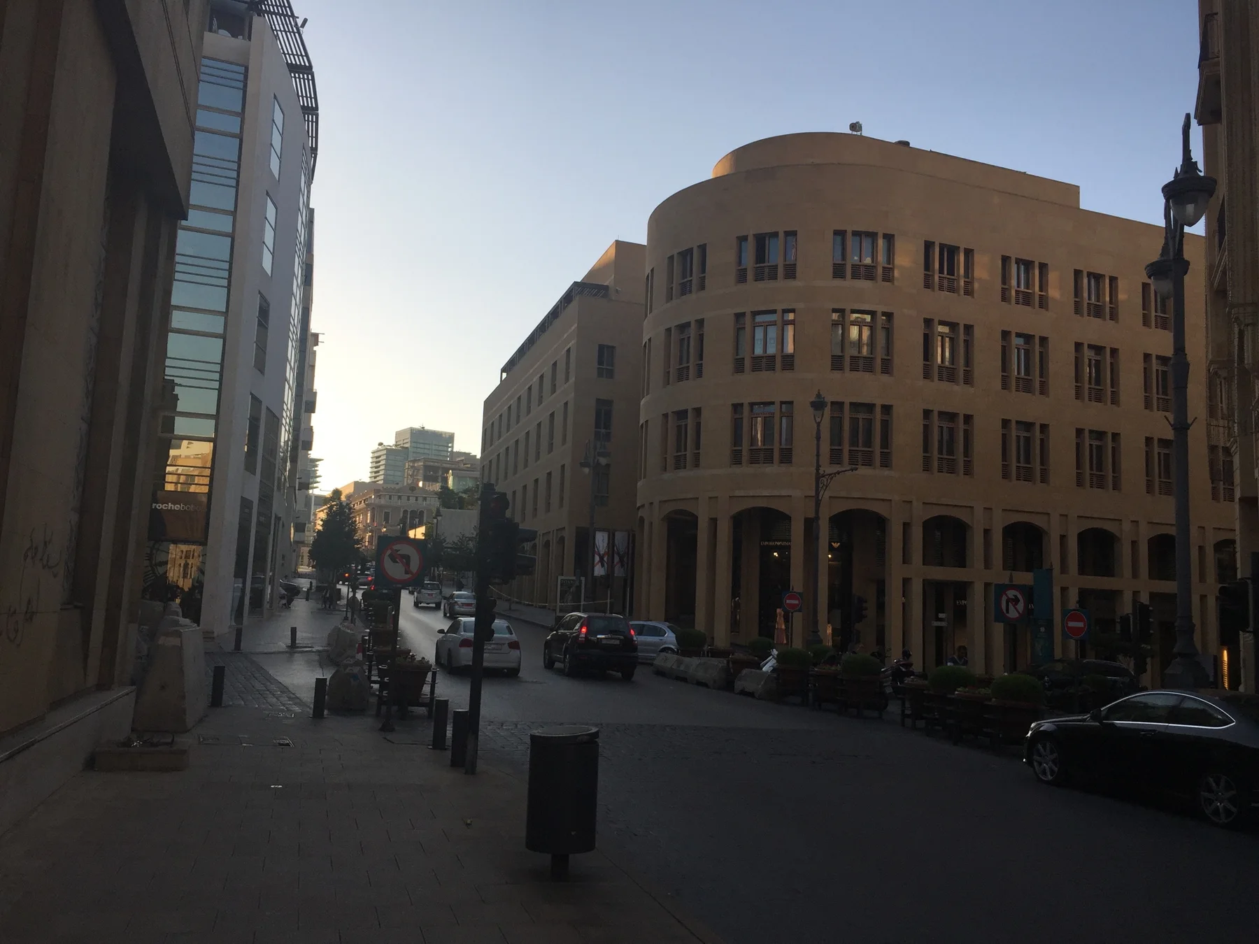 A quiet street runs beside a curved ochre building and pale stone blocks in low sunlight.
