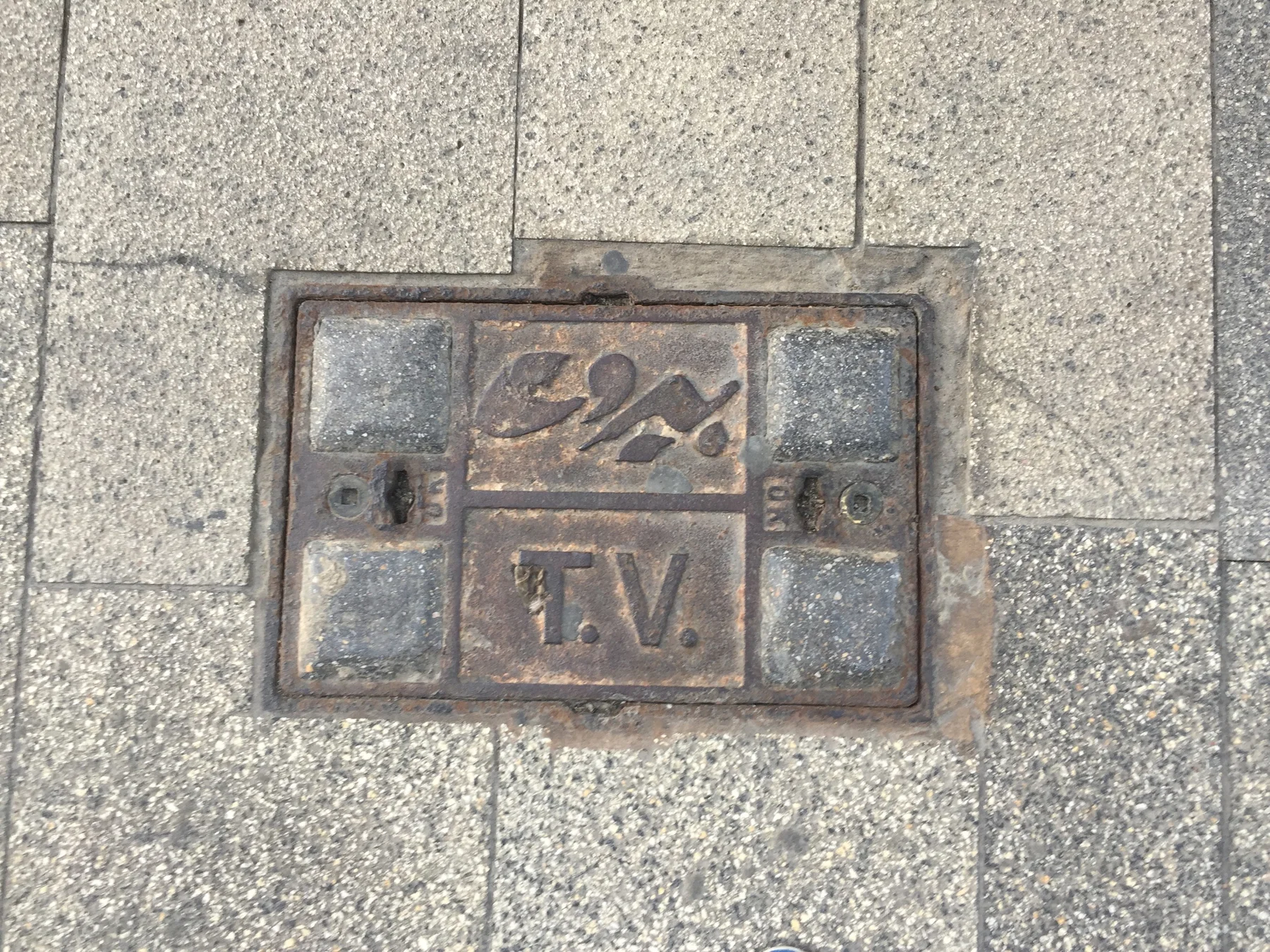 A small square metal service cover with lettering is set into a grid of stone paving.