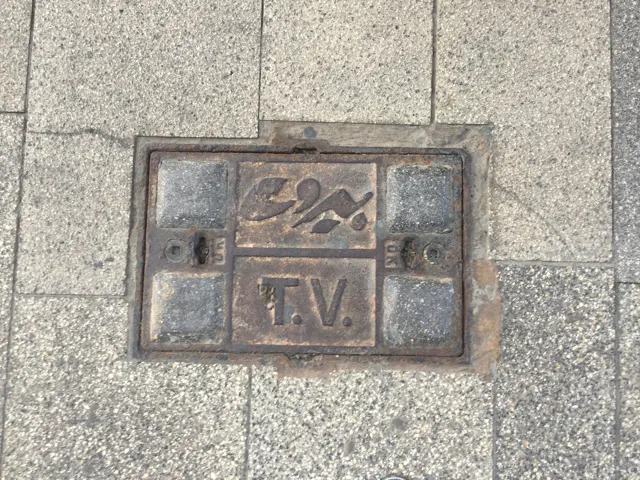 A small square metal service cover with lettering is set into a grid of stone paving.