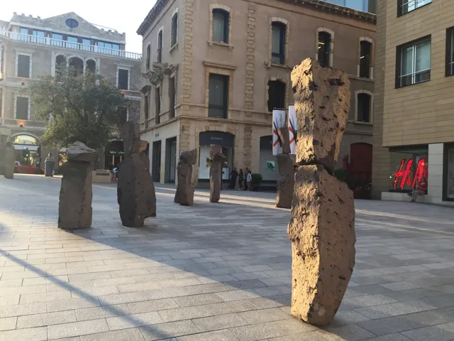 Several tall rough-textured sculptures stand in a paved square surrounded by buildings.