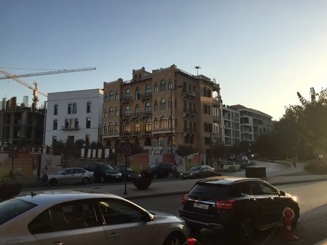 Cars pass a weathered corner building beside newer structures in low evening light.