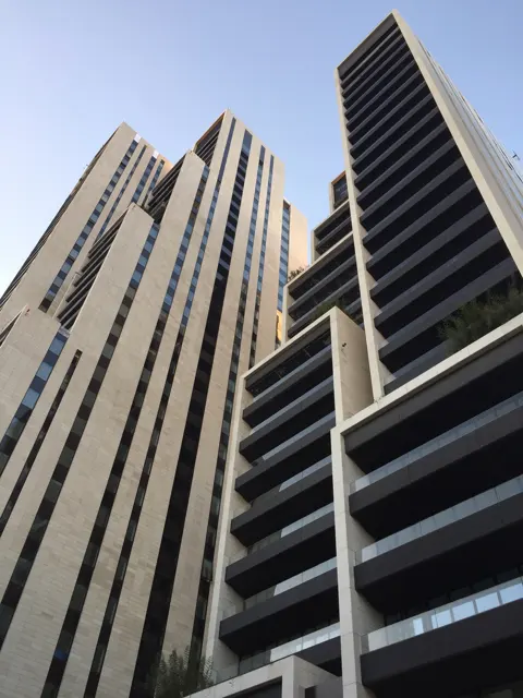 An upward portrait view follows tall ribbed building facades and stacked balconies against a blue sky.