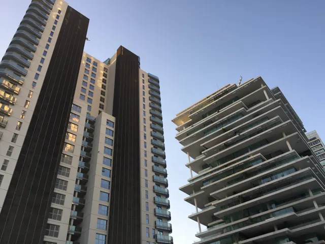 A tall ribbed tower and a building with wide horizontal balconies frame a patch of blue sky.