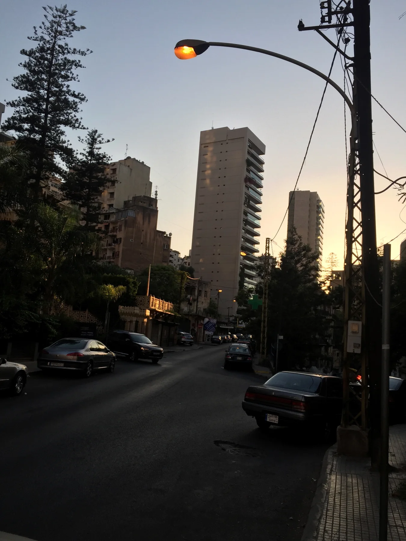 Parked cars line a shaded street beneath a lit lamp, with high-rise silhouettes against the fading sky.