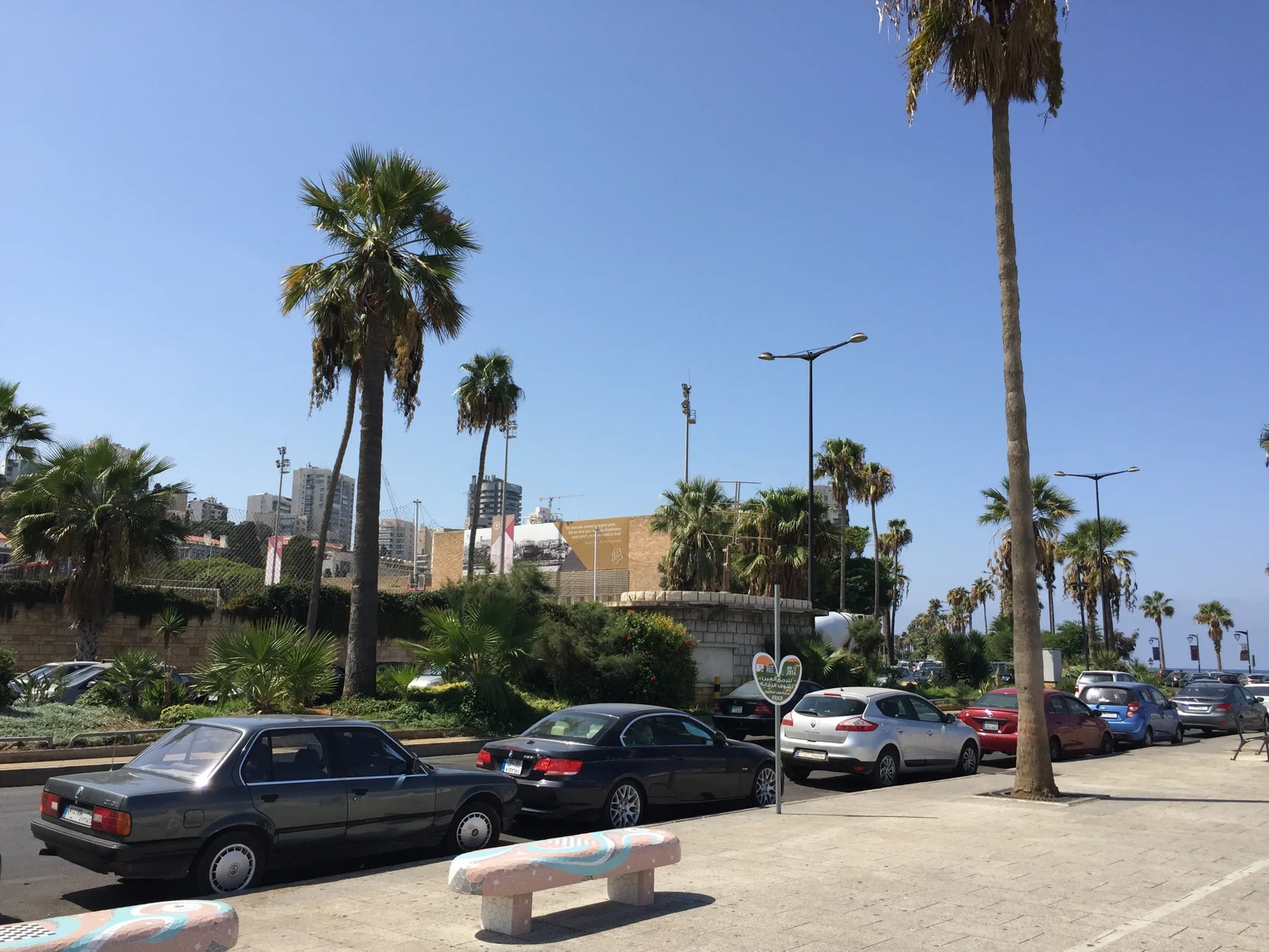 Cars line a road beside palms, low coastal buildings and a paved promenade under blue skies.