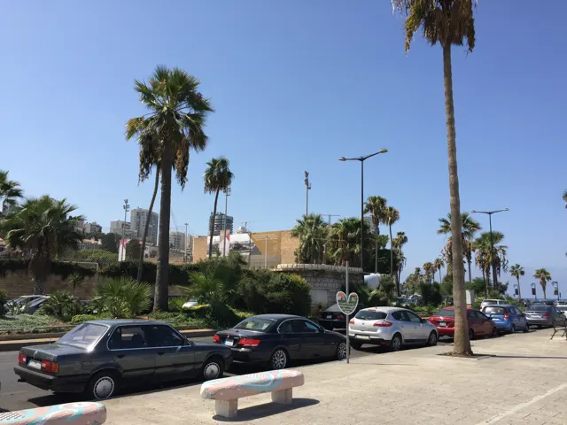 Cars line a road beside palms, low coastal buildings and a paved promenade under blue skies.