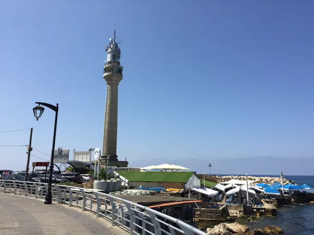 A tall pale tower rises beside a waterfront walkway and low buildings at the edge of the sea.