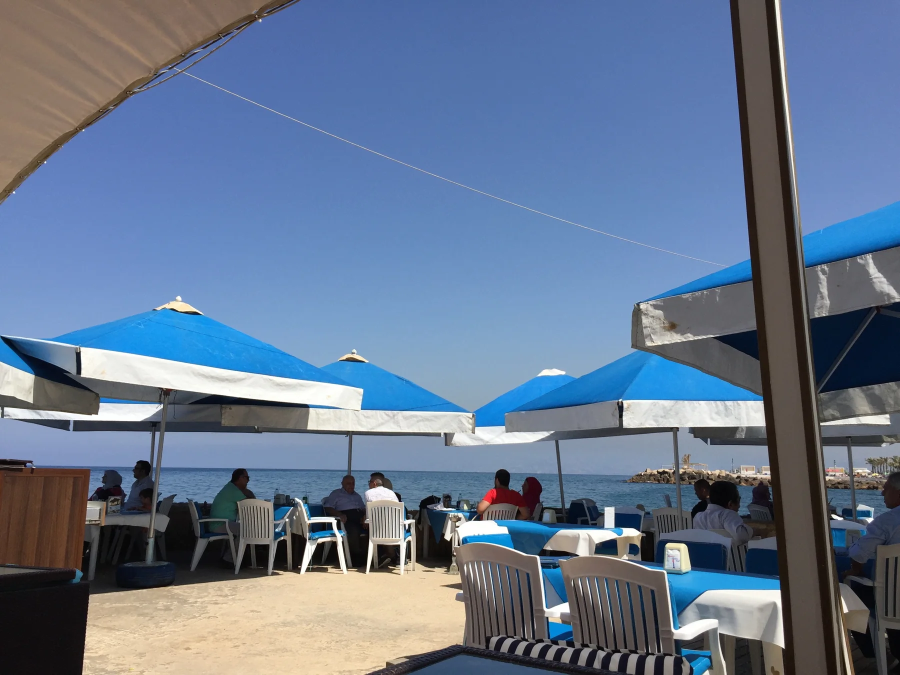 Blue-and-white umbrellas shade outdoor tables and chairs at a seaside terrace.