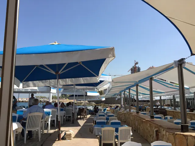 Rows of tables and chairs sit beneath blue umbrellas and a pale canopy beside the water.